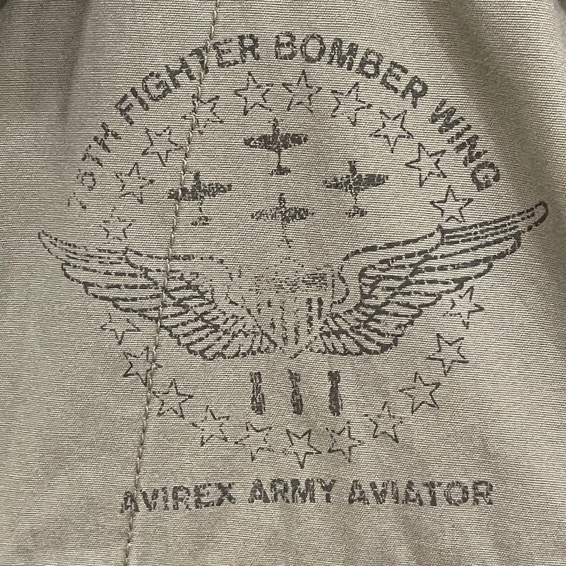 K&ouml;nnte beinhalten: Nahaufnahme eines beigefarbenen Stoffes mit einem Used-Print. Der Druck zeigt ein kreisf&ouml;rmiges Design mit Sternen, Fl&uuml;geln und dem Text "8th Fighter Bomber Wing" und "Avirex Army Aviator".