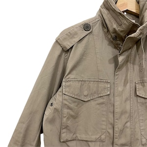 K&ouml;nnte beinhalten: Eine milit&auml;risch anmutende Jacke in Beige mit hohem Kragen und mehreren Taschen. Die Jacke hat Knopfverschl&uuml;sse, Schulterklappen und einen Rei&szlig;verschluss am Kragen. Der Stoff scheint strapazierf&auml;hig zu sein, m&ouml;glicherweise Baumwolle oder eine Baumwollmischung.