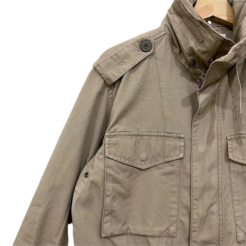 K&ouml;nnte beinhalten: Eine milit&auml;risch anmutende Jacke in Beige mit hohem Kragen und mehreren Taschen. Die Jacke hat Knopfverschl&uuml;sse, Schulterklappen und einen Rei&szlig;verschluss am Kragen. Der Stoff scheint strapazierf&auml;hig zu sein, m&ouml;glicherweise Baumwolle oder eine Baumwollmischung.