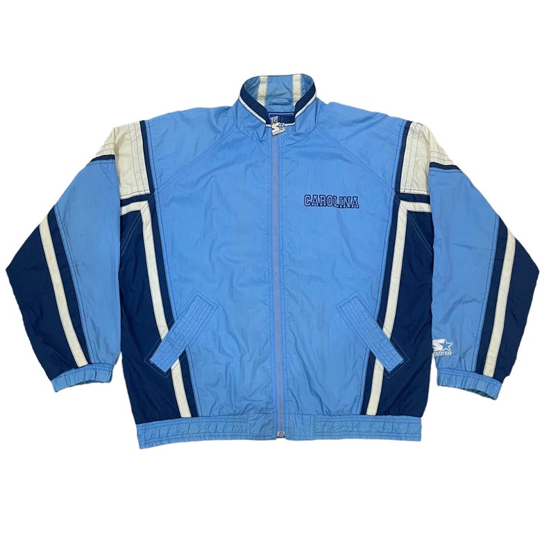 May include: Light blue and navy blue Starter jacket with white accents. The jacket features the word "CAROLINA" embroidered across the chest. It has a full zipper, two front pockets, and a stand-up collar. The jacket is a vintage style.