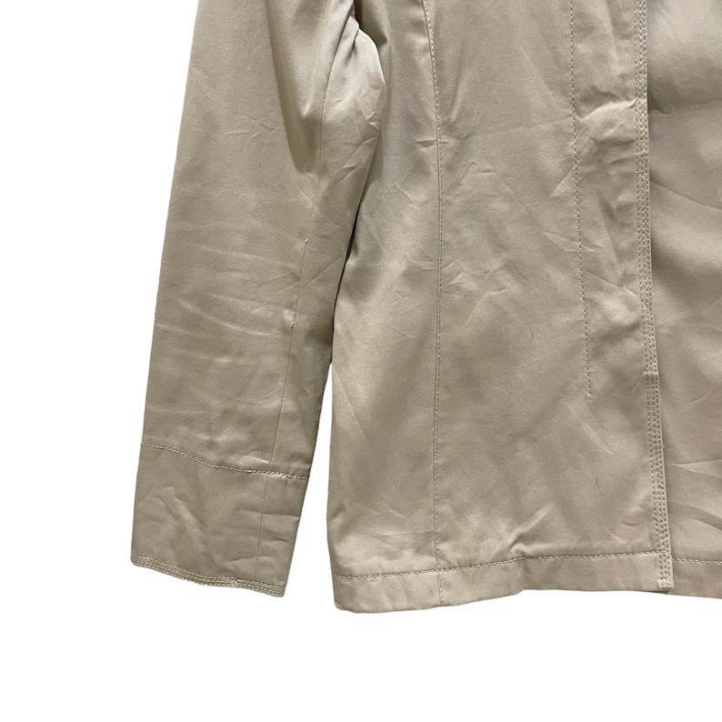 May include: Close-up of a tan jacket. The jacket has a simple design with a tailored fit. The sleeve is visible, showing a cuff detail. The fabric has a subtle sheen, and the stitching is neat and precise. The jacket appears to be made of a lightweight material.