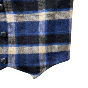 May include: A sleeveless vest featuring a plaid pattern in shades of navy blue, bright blue, and beige. The garment has a pointed hemline and is fastened with three round, black, textured buttons along the left side of the front panel.