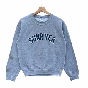 May include: A heather gray crewneck sweatshirt featuring the word SUNRIVER in dark blue block letters with a white outline across the chest. The long-sleeved garment has a ribbed collar, cuffs, and hem, and displays visible dark stains on the left sleeve.