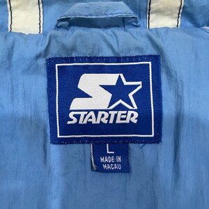 May include: Close-up of a light blue Starter jacket tag. The tag is a blue rectangle with the white Starter logo and star. The tag also has the size "L" and "MADE IN MACAU" printed on it. The jacket has white stripes.