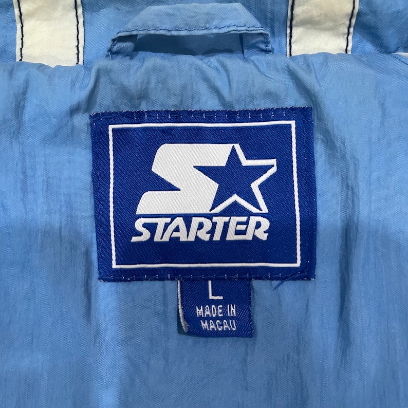 May include: Close-up of a light blue Starter jacket tag. The tag is a blue rectangle with the white Starter logo and star. The tag also has the size "L" and "MADE IN MACAU" printed on it. The jacket has white stripes.