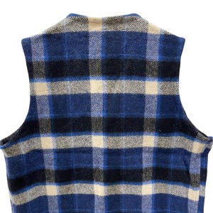 May include: A sleeveless vest featuring a plaid pattern in shades of navy blue, royal blue, black, and cream. The garment has a round neckline and a straight hem, showcasing a textured fabric construction with a consistent grid-like design throughout.