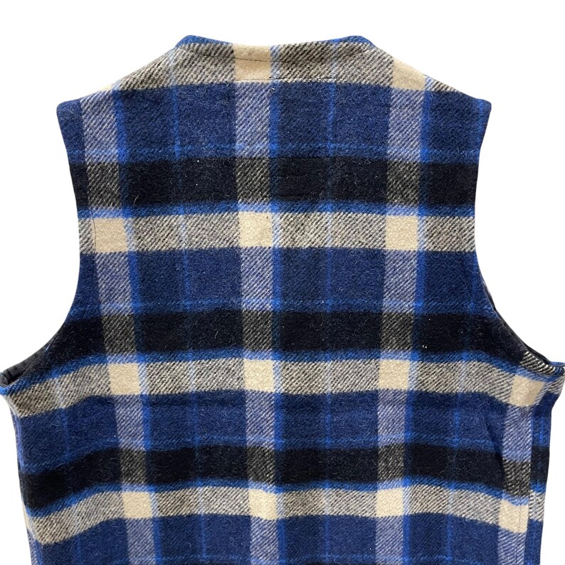 May include: A sleeveless vest featuring a plaid pattern in shades of navy blue, royal blue, black, and cream. The garment has a round neckline and a straight hem, showcasing a textured fabric construction with a consistent grid-like design throughout.