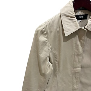 May include: A beige IN&Eacute;D trench coat. The coat features a classic collar, a button closure, and a tailored fit. The brand name IN&Eacute;D is visible on the label. The coat is made of a smooth, lightweight fabric, suitable for transitional weather.