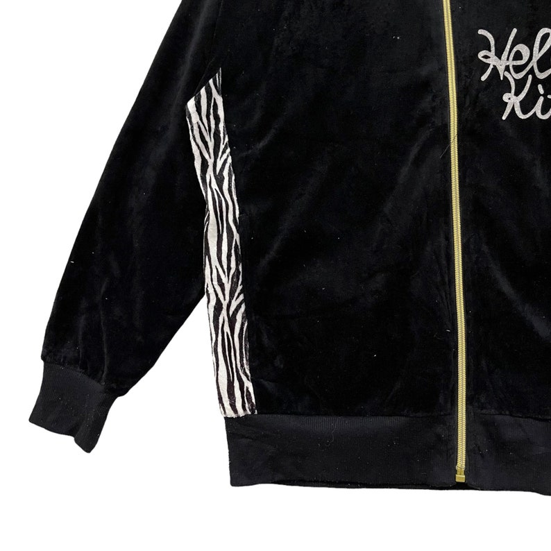 May include: Black velour jacket with a zipper closure and a zebra print panel on the side. The jacket has a gold zipper and the text "Hello Kitty" is embroidered on the right side.