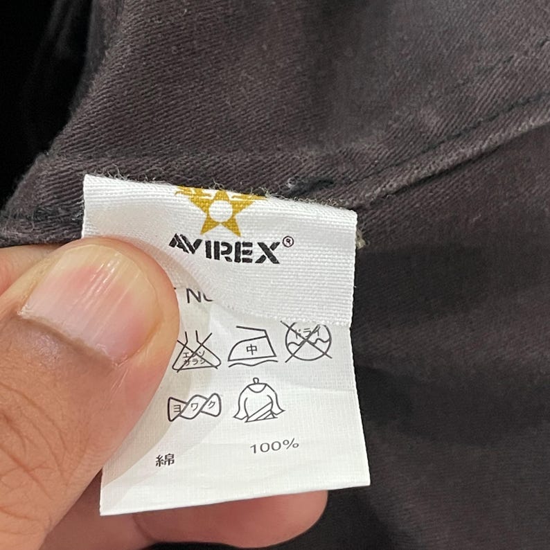 K&ouml;nnte beinhalten: Ein wei&szlig;es Textil-Pflegeetikett an einem dunkelbraunen Kleidungsst&uuml;ck. Das Etikett zeigt das Avirex-Logo mit einem goldenen Stern, den Text 100% Baumwolle sowie verschiedene Pflegesymbole f&uuml;r das Waschen, B&uuml;geln und Trocknen der Kleidung.