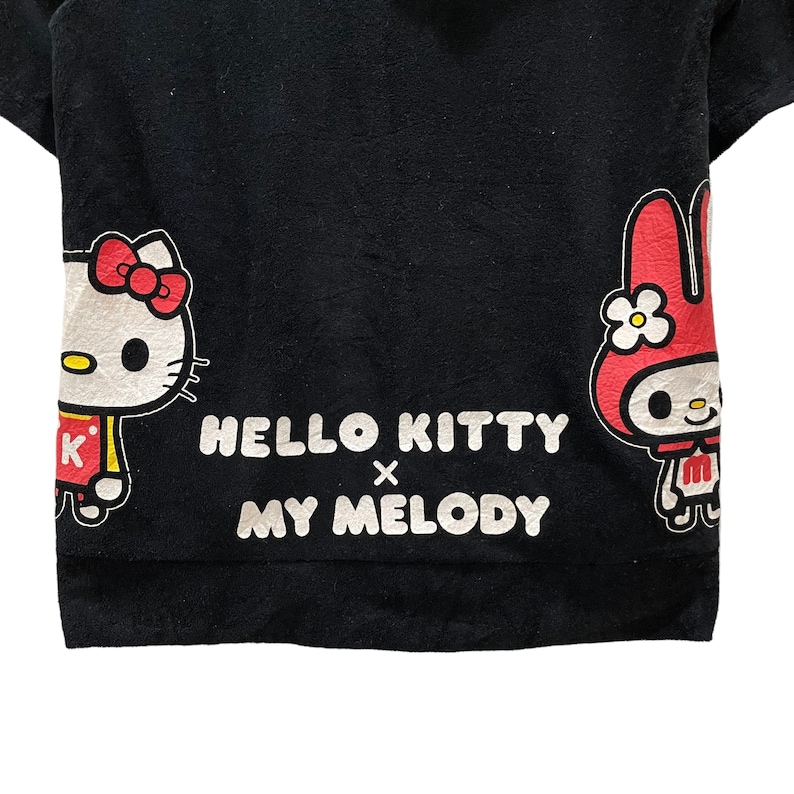 May include: Black t-shirt with Hello Kitty and My Melody characters. Hello Kitty is on the left with a red bow, and My Melody is on the right. The text "HELLO KITTY x MY MELODY" is printed in white across the centre.