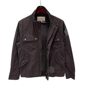 K&ouml;nnte beinhalten: Eine dunkelbraune Feldjacke mit Stehkragen, Frontrei&szlig;verschluss mit Druckkn&ouml;pfen und vier Vordertaschen. Die Jacke ist mit wei&szlig;em Text auf der Brust und dem &Auml;rmel bedruckt und verf&uuml;gt &uuml;ber ein Markenetikett im inneren Nackenbereich.