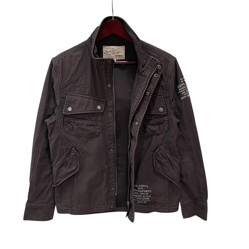 K&ouml;nnte beinhalten: Eine dunkelbraune Feldjacke mit Stehkragen, Frontrei&szlig;verschluss mit Druckkn&ouml;pfen und vier Vordertaschen. Die Jacke ist mit wei&szlig;em Text auf der Brust und dem &Auml;rmel bedruckt und verf&uuml;gt &uuml;ber ein Markenetikett im inneren Nackenbereich.