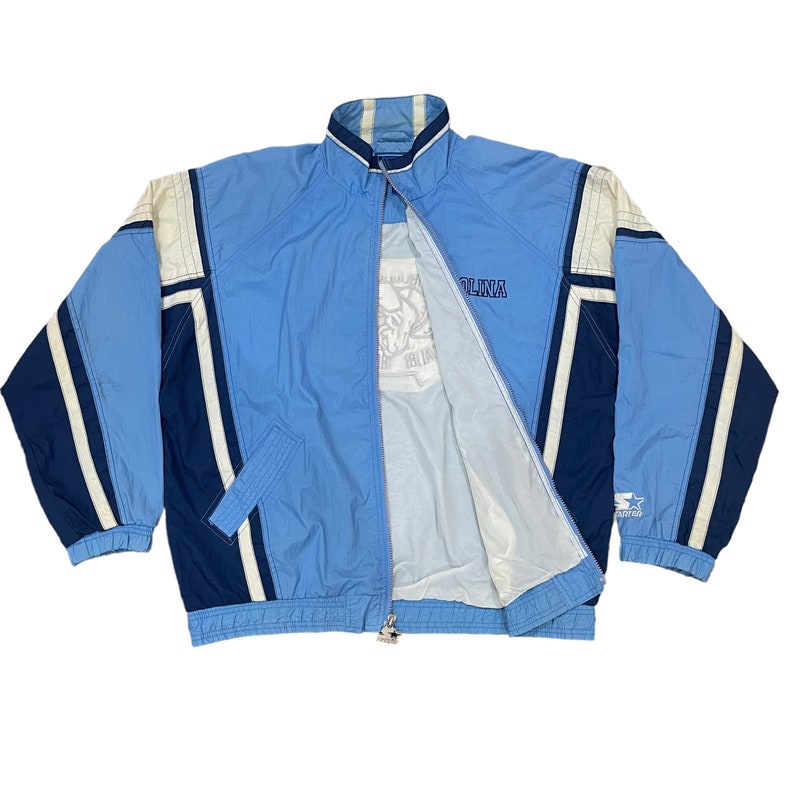 May include: Light blue and navy blue Starter jacket with white accents. The jacket has a full zipper, a stand-up collar, and the word "CAROLINA" embroidered on the chest. The Starter logo is visible on the sleeve.