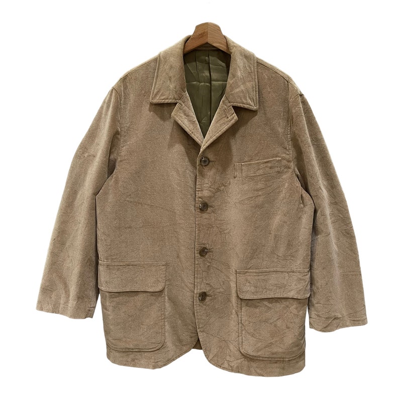Puede incluir: Un blazer de pana color beige con un dise&ntilde;o cl&aacute;sico. La chaqueta presenta solapas con muescas, cierre de cuatro botones y dos bolsillos delanteros. El forro interior es de color verde oliva en contraste. El blazer est&aacute; colgado de una percha de madera.