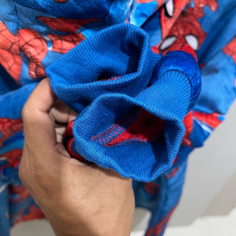 Puede incluir: Primer plano de un mono azul y rojo con el tema de Spider-Man. Los pu&ntilde;os son de color azul liso con costuras rojas. El mono tiene un patr&oacute;n repetido de la cara de Spider-Man.