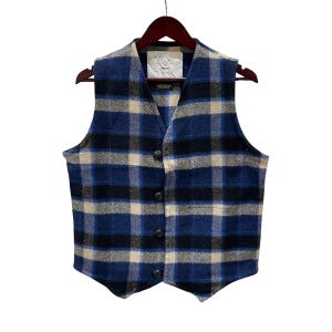 May include: A sleeveless vest featuring a blue, black, and cream plaid pattern. The garment has a V-neckline, four dark circular buttons down the front, and a pointed hemline. A white rectangular label is visible at the inner neckline.