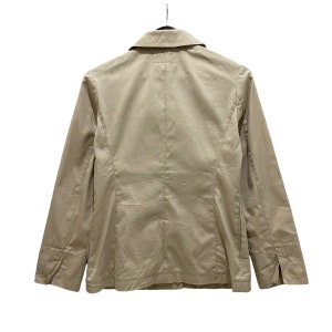 May include: A tan blazer with a classic collar and long sleeves, hanging against a white background. The jacket features a button closure and a tailored fit, showcasing a minimalist design. The back view highlights the structured seams and overall silhouette.