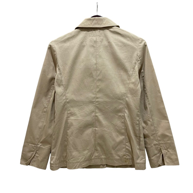 May include: A tan blazer with a classic collar and long sleeves, hanging against a white background. The jacket features a button closure and a tailored fit, showcasing a minimalist design. The back view highlights the structured seams and overall silhouette.