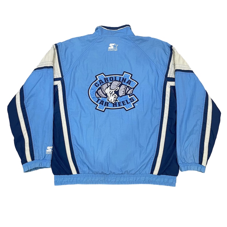 May include: Light blue Carolina Tar Heels Starter jacket. The jacket features a full zip, a stand-up collar, and the team logo on the back. White and navy blue accents are on the sleeves and sides. The Starter logo is visible on the chest and sleeve.