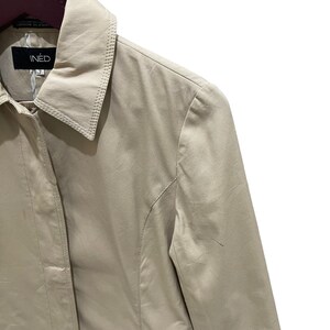 May include: A beige INED jacket with a collared neckline. The jacket has a button closure and a tailored fit. The brand name "INED" is visible on a black label. The jacket is made of a smooth, possibly satin-like material.