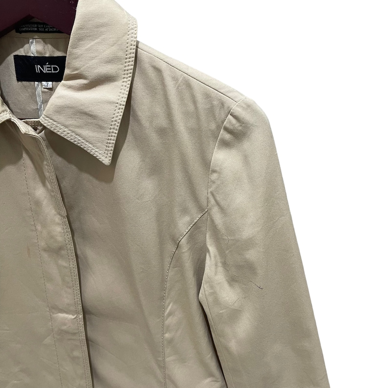 May include: A beige INED jacket with a collared neckline. The jacket has a button closure and a tailored fit. The brand name "INED" is visible on a black label. The jacket is made of a smooth, possibly satin-like material.