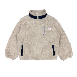 May include: A beige fleece jacket with a high collar and a full-length zip. The jacket features navy blue detailing on the collar, pockets, and a small patch. The jacket is made of a soft, textured material.