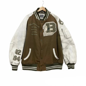 May include: A brown and white varsity jacket with a large "B" on the front. The sleeves are white leather and the body is brown with white stripes. The jacket has a zipper and button closure.