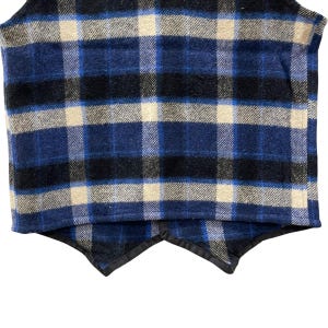 May include: A sleeveless vest featuring a plaid pattern in shades of navy blue, black, and cream. The garment has a textured fabric finish and a hemline with two pointed front panels finished with a black trim along the bottom edges.