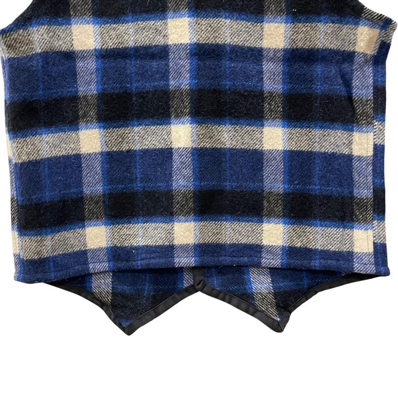 May include: A sleeveless vest featuring a plaid pattern in shades of navy blue, black, and cream. The garment has a textured fabric finish and a hemline with two pointed front panels finished with a black trim along the bottom edges.