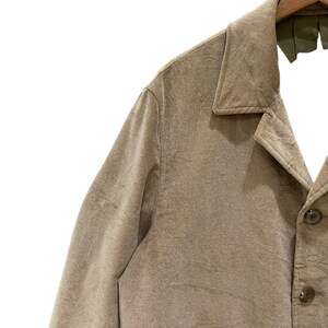 Puede incluir: Primer plano de una chaqueta de pana color beige con cuello cl&aacute;sico y cierre de botones. La chaqueta est&aacute; colgada de una percha de madera. La tela tiene un aspecto texturizado.