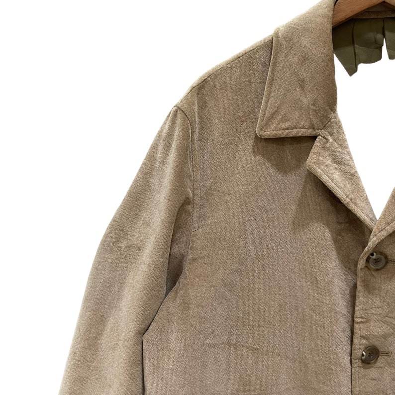 Puede incluir: Primer plano de una chaqueta de pana color beige con cuello cl&aacute;sico y cierre de botones. La chaqueta est&aacute; colgada de una percha de madera. La tela tiene un aspecto texturizado.