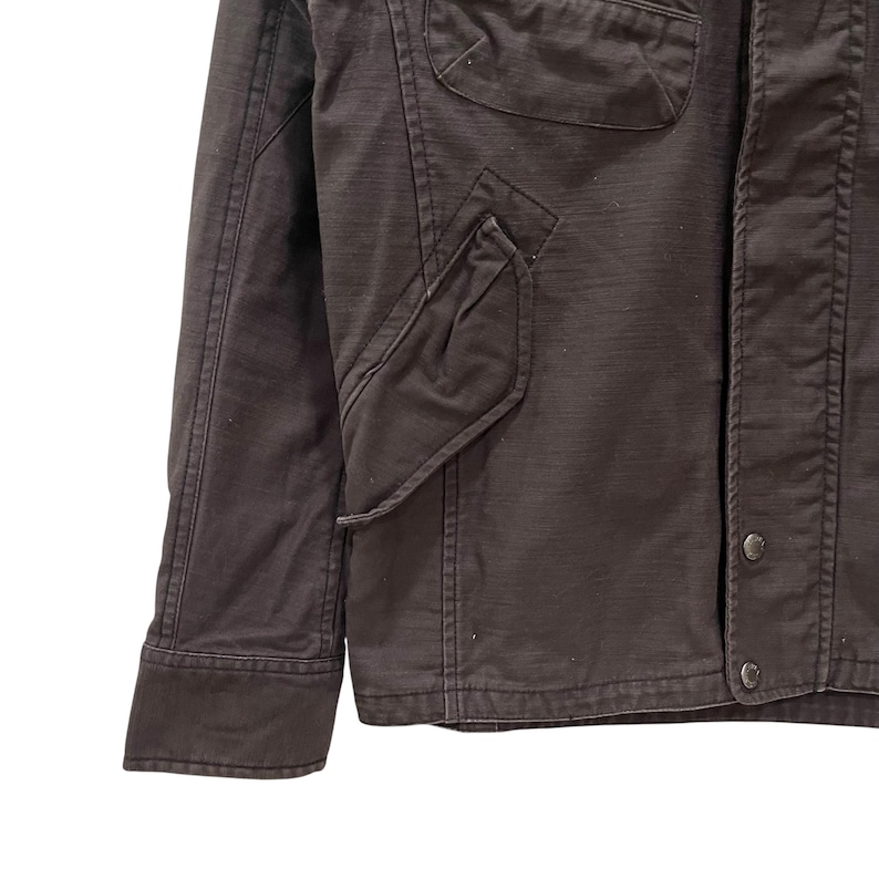 K&ouml;nnte beinhalten: Eine dunkelbraune Jacke mit strukturiertem Kragen, einer Knopfleiste mit sichtbaren Metalldruckkn&ouml;pfen und zwei markanten Taschen im Brustbereich. Das Kleidungsst&uuml;ck ist mit verst&auml;rkten N&auml;hten entlang der &Auml;rmel und Partien versehen.