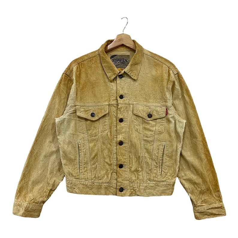 May include: A tan corduroy jacket with black buttons and two chest pockets. The jacket has a classic design with a collar and long sleeves. A small red tag is visible on the left side. The brand name "EDWIN" is visible on the label.