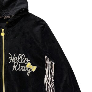 May include: Black velour jacket with a zipper closure, a hood, and a Hello Kitty logo with a yellow bow on the front. The jacket has a zebra print stripe on the right side.