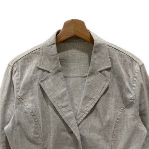 Puede incluir: Un blazer gris claro con solapa de muesca y cierre de un solo bot&oacute;n. La chaqueta est&aacute; hecha de una tela texturizada y se muestra en una percha de madera. El blazer tiene un dise&ntilde;o cl&aacute;sico y entallado.