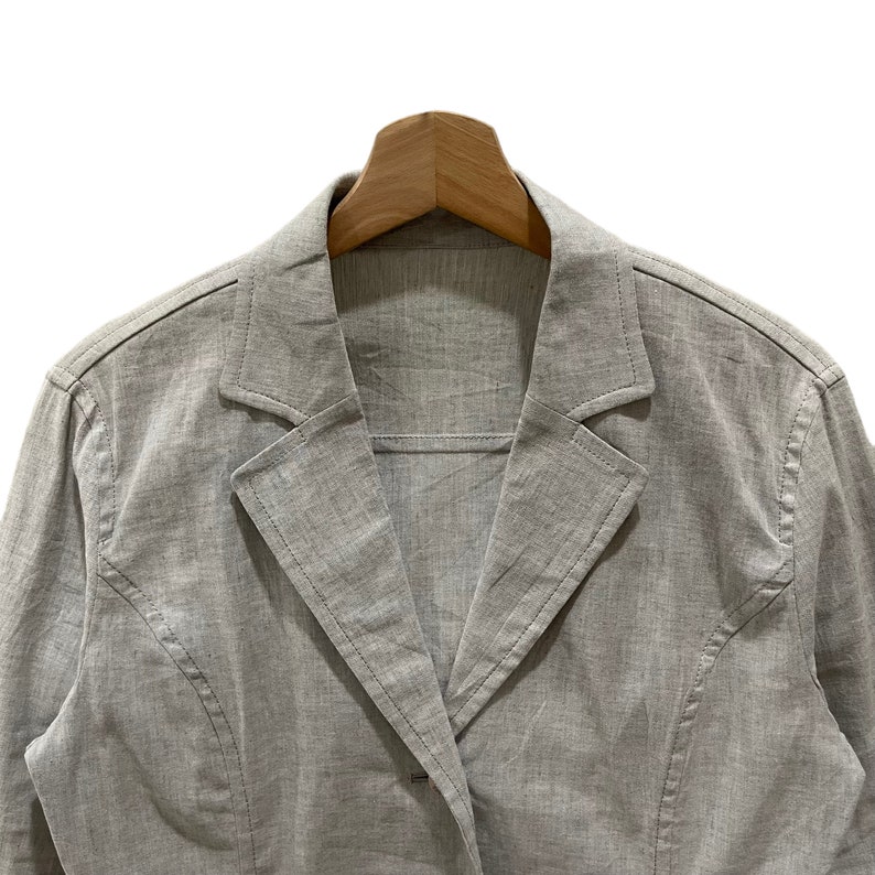 Puede incluir: Un blazer gris claro con solapa de muesca y cierre de un solo bot&oacute;n. La chaqueta est&aacute; hecha de una tela texturizada y se muestra en una percha de madera. El blazer tiene un dise&ntilde;o cl&aacute;sico y entallado.