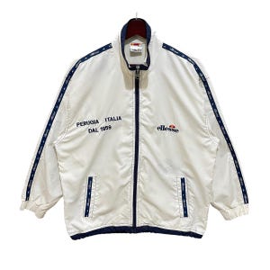 May include: White zip-up jacket with navy blue trim and accents. The jacket features the text "PERUGIA ITALIA DAL 1959" and the brand name "ellesse". The jacket has a stand-up collar and two front pockets.
