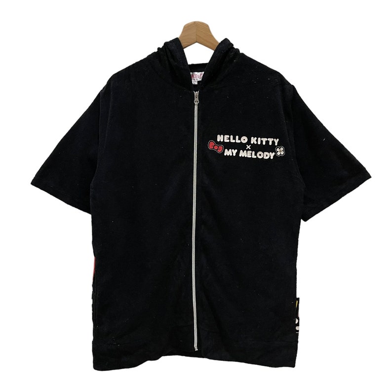 May include: Black hooded zip-up short-sleeved shirt. The shirt features the text "HELLO KITTY x MY MELODY" in white and red. The shirt is hanging on a wooden hanger.