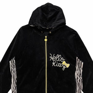 May include: Black hooded jacket with a zipper closure and a zebra print on the sleeves. The jacket has a Hello Kitty logo with a yellow bow on the front.