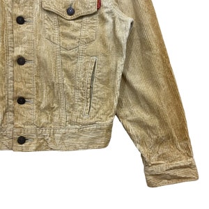 May include: Close-up of a tan corduroy jacket with a button-down front, chest pocket, and side pocket. The jacket has dark brown buttons and a small red tag on the chest. The corduroy fabric has vertical ridges.