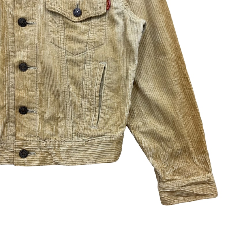 May include: Close-up of a tan corduroy jacket with a button-down front, chest pocket, and side pocket. The jacket has dark brown buttons and a small red tag on the chest. The corduroy fabric has vertical ridges.
