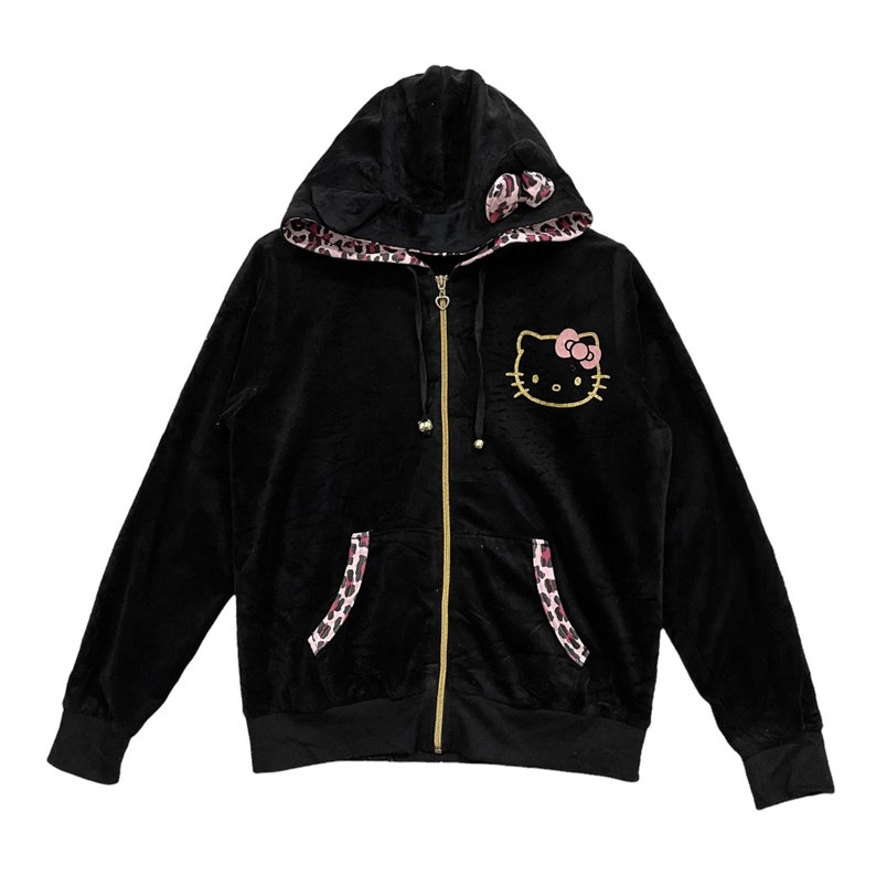 Pode incluir: Casaco de moletom preto de veludo com z&iacute;per dourado e um gr&aacute;fico de Hello Kitty na frente. O capuz &eacute; forrado com uma estampa de leopardo rosa e preto.