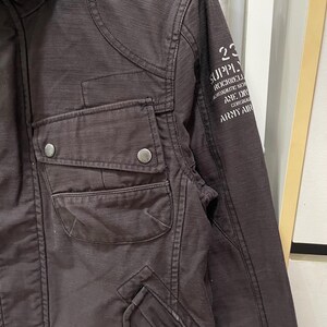 K&ouml;nnte beinhalten: Eine dunkelbraune Jacke mit einer Brusttasche und zwei silberfarbenen Metalldruckkn&ouml;pfen. Auf dem &Auml;rmel befindet sich ein wei&szlig;er Aufdruck mit dem Text 23 SUPPLY ROCKWELL AERONAUTIC SER AME DIV CORONAD ARMY AIR. Der Stoff hat eine sichtbare Webstruktur.