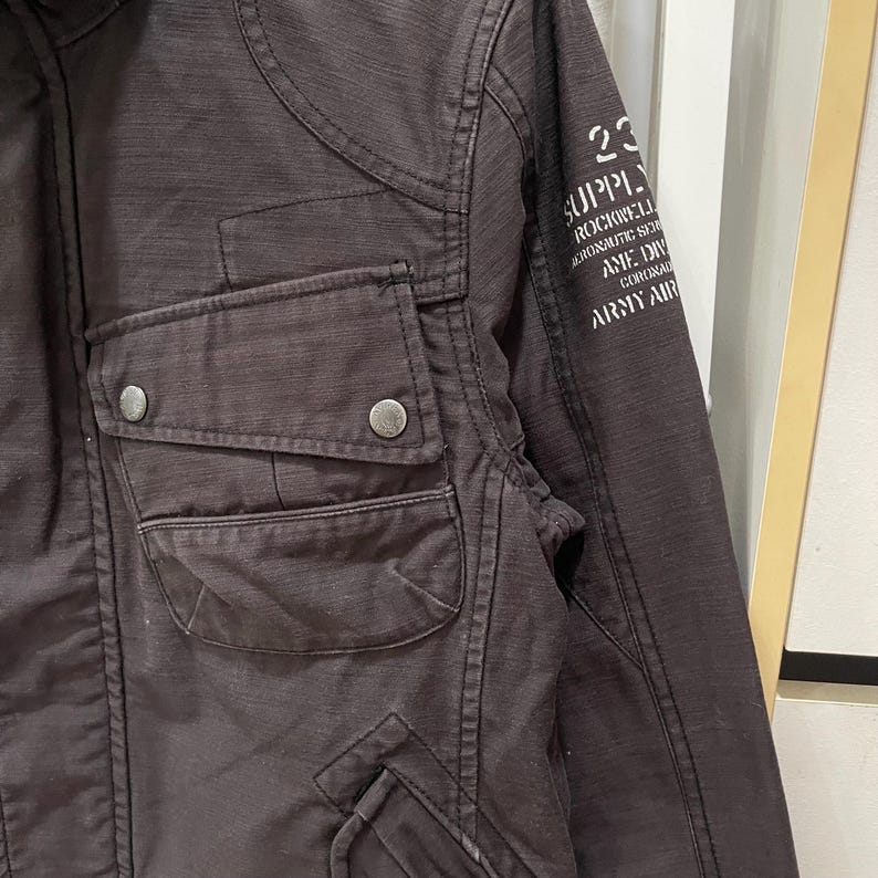 K&ouml;nnte beinhalten: Eine dunkelbraune Jacke mit einer Brusttasche und zwei silberfarbenen Metalldruckkn&ouml;pfen. Auf dem &Auml;rmel befindet sich ein wei&szlig;er Aufdruck mit dem Text 23 SUPPLY ROCKWELL AERONAUTIC SER AME DIV CORONAD ARMY AIR. Der Stoff hat eine sichtbare Webstruktur.