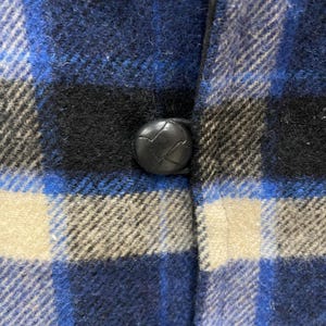 May include: A blue, black, and off-white plaid flannel shirt featuring a single round, black, textured button centered on the placket. The fabric has a soft, brushed appearance with a classic woven check pattern throughout the garment.