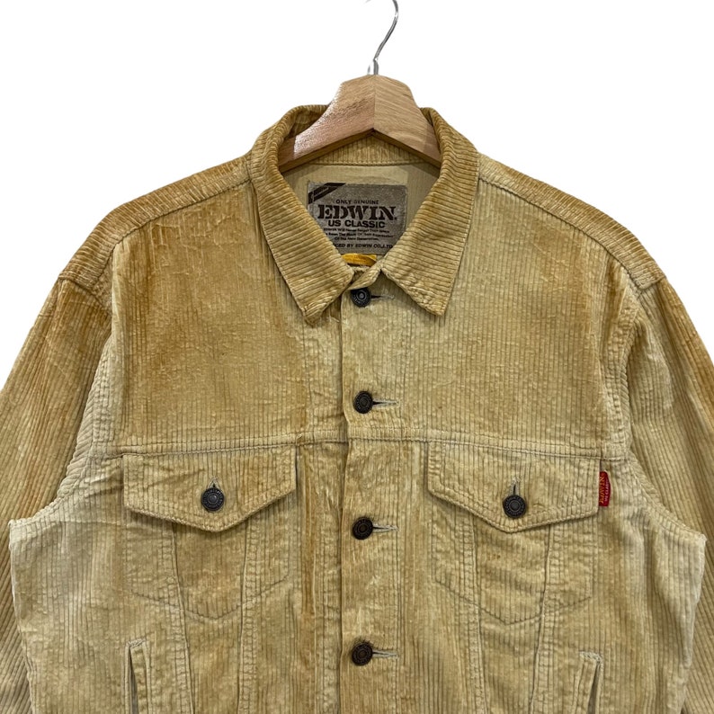 May include: A tan corduroy jacket with a button-up front, two chest pockets, and two side pockets. The jacket has a classic collar and a small red tag on the right side. The brand name "EDWIN US CLASSIC" is visible on the label.