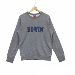 May include: A gray crew neck sweatshirt with the word "EDWIN" embroidered in navy blue across the chest. The sweatshirt is hanging on a wooden hanger against a white background. The brand tag is visible on the back of the neck.