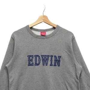 May include: A gray Edwin sweatshirt with the brand name embroidered in navy blue across the chest. The sweatshirt is hanging on a wooden hanger against a white background. A small red tag with the brand name is visible.