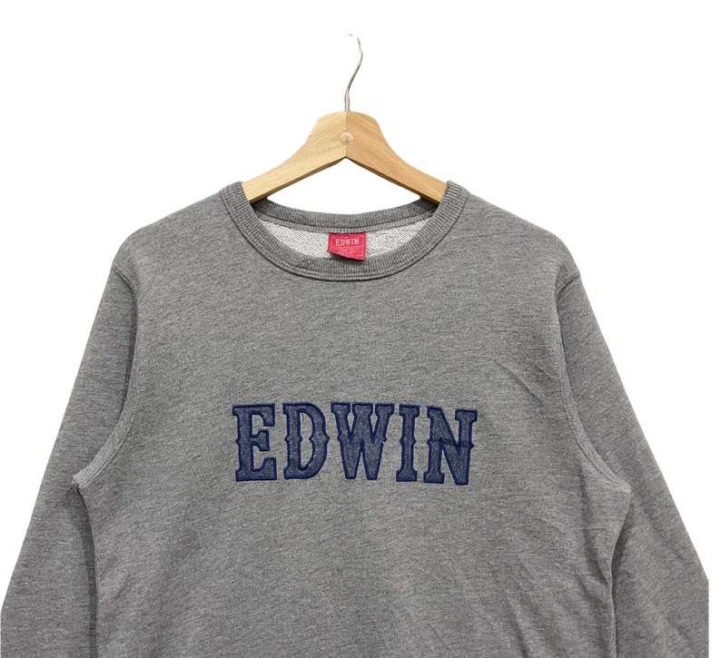 May include: A gray Edwin sweatshirt with the brand name embroidered in navy blue across the chest. The sweatshirt is hanging on a wooden hanger against a white background. A small red tag with the brand name is visible.